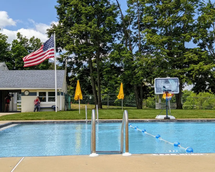 Main Pool with a steel ladder, lane line and yellow umbrellas, flag and basketball hoop beyond